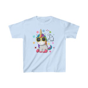 Könnte beinhalten: Hellblaues T-Shirt mit einem Comic-Einhorn, das eine Sonnenbrille trägt und eine Regenbogenmähne hat. Das Einhorn ist von bunten Blumen umgeben und der Text "Be Cool" ist auf das T-Shirt gedruckt.