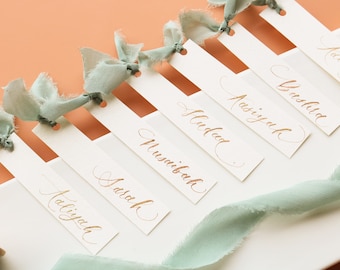 Può includere: Un set di segnaposto bianchi con nomi in calligrafia dorata, legati con un nastro blu chiaro. I nomi sono: "Talliyah", "Sarah", "Musnibah", "Huddad", "Aasiyah", "Yreshia" e "Aisha".
