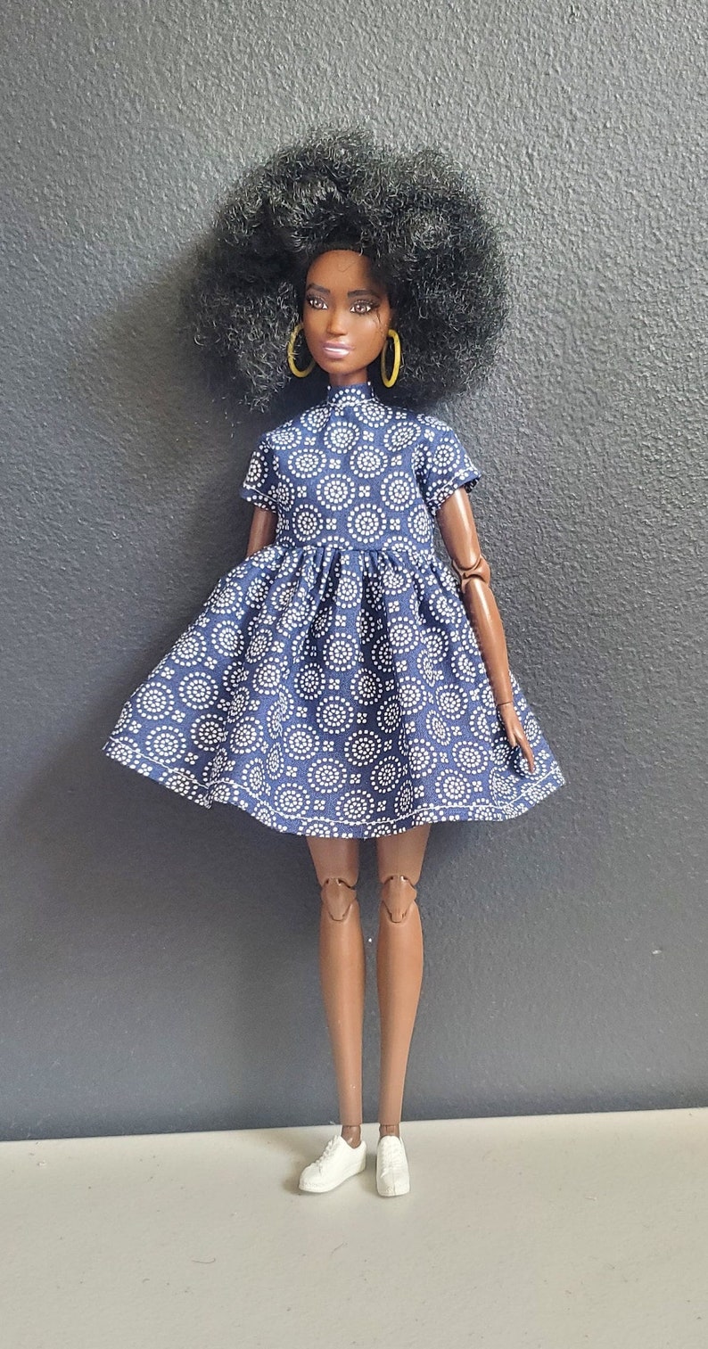 K&ouml;nnte beinhalten: Eine Puppe mit dunkelbrauner Haut und einer gro&szlig;en Afrofrisur tr&auml;gt ein blaues Kleid mit einem wei&szlig;en geometrischen Muster. Das Kleid hat kurze &Auml;rmel und einen runden Ausschnitt. Die Puppe tr&auml;gt wei&szlig;e Turnschuhe.