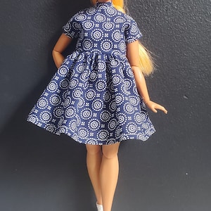 K&ouml;nnte beinhalten: Eine Puppe, die ein blau-wei&szlig; gemustertes Kleid mit wei&szlig;en Turnschuhen tr&auml;gt.