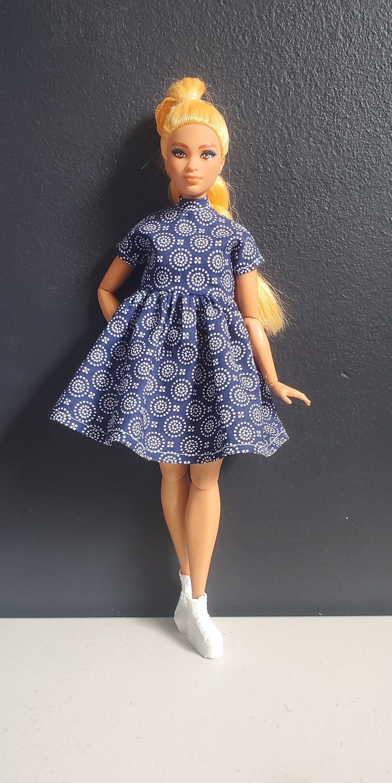 K&ouml;nnte beinhalten: Eine Puppe, die ein blau-wei&szlig; gemustertes Kleid mit wei&szlig;en Turnschuhen tr&auml;gt.