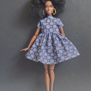 K&ouml;nnte beinhalten: Eine Puppe tr&auml;gt ein blau-wei&szlig; gemustertes Kleid mit hohem Kragen und kurzen &Auml;rmeln. Die Puppe hat braune Haut, lockiges schwarzes Haar und tr&auml;gt gelbe Creolen. Die Puppe steht auf einer grauen Oberfl&auml;che.