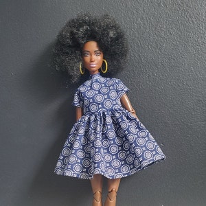 K&ouml;nnte beinhalten: Eine schwarze Puppe mit dunkelbraunen Augen und einem dunkelbraunen Afro, die ein blau-wei&szlig; gemustertes Kleid mit gelben Ohrringen tr&auml;gt.