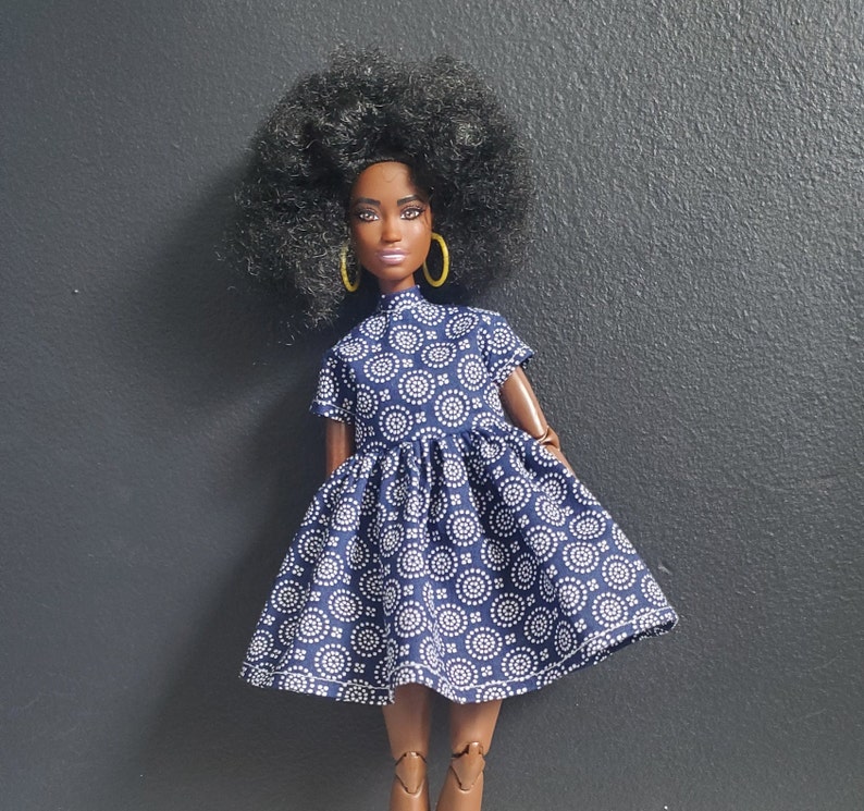 K&ouml;nnte beinhalten: Eine schwarze Puppe mit dunkelbraunen Augen und einem dunkelbraunen Afro, die ein blau-wei&szlig; gemustertes Kleid mit gelben Ohrringen tr&auml;gt.