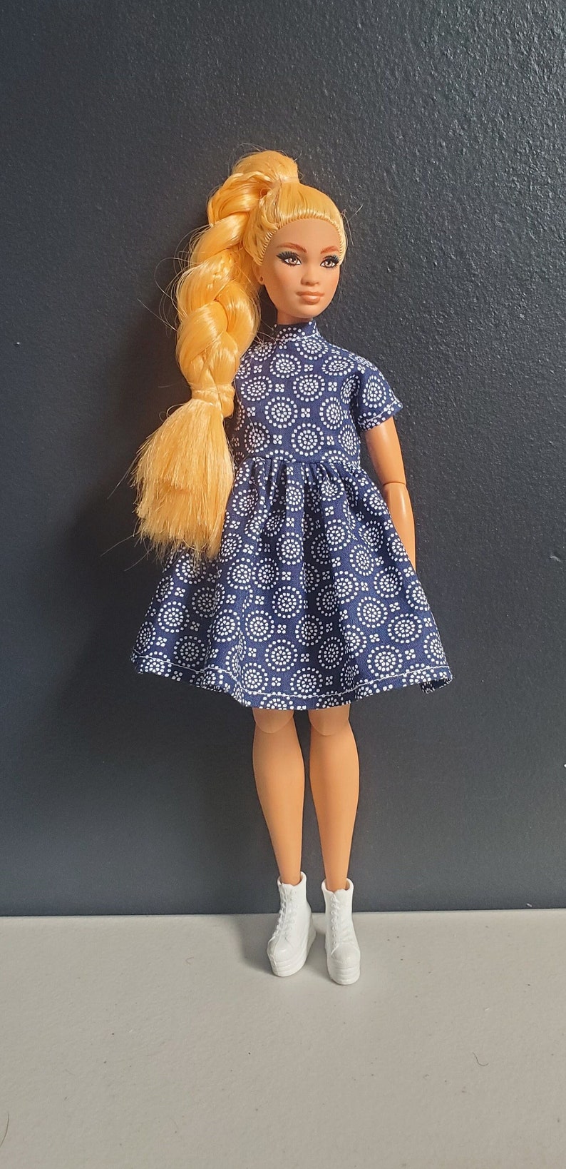 K&ouml;nnte beinhalten: Eine blonde Puppe tr&auml;gt ein blaues und wei&szlig; gemustertes Kleid mit hohem Kragen und kurzen &Auml;rmeln. Die Puppe tr&auml;gt wei&szlig;e Turnschuhe.