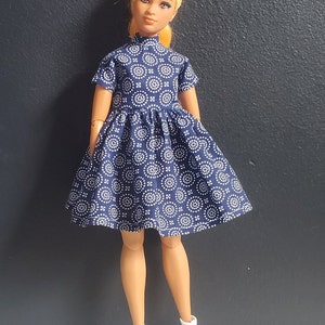 K&ouml;nnte beinhalten: Eine Puppe tr&auml;gt ein blaues Kleid mit einem wei&szlig;en geometrischen Muster. Das Kleid hat einen hohen Kragen und kurze &Auml;rmel. Die Puppe tr&auml;gt wei&szlig;e Schuhe.