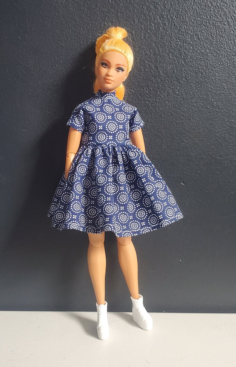 K&ouml;nnte beinhalten: Eine Puppe tr&auml;gt ein blaues Kleid mit einem wei&szlig;en geometrischen Muster. Das Kleid hat einen hohen Kragen und kurze &Auml;rmel. Die Puppe tr&auml;gt wei&szlig;e Schuhe.