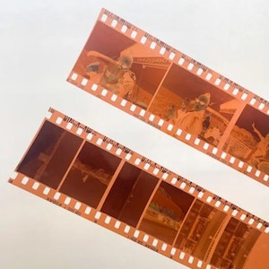 Puede incluir: Dos tiras de negativos de película teñidos de naranja con múltiples fotogramas. Las tiras de película tienen orificios de sprocket a lo largo de los bordes y contienen imágenes de personas y edificios. El texto "KODAK" y "GB 200-7" es visible en la película.