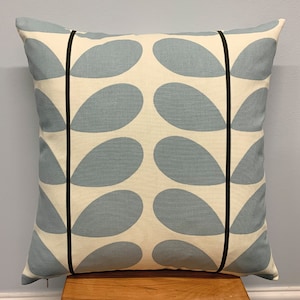 May include: Square throw pillow with a light blue teardrop pattern on a cream background. The pillow has black vertical stripes and a zipper closure. The pillow is placed on a wooden surface.