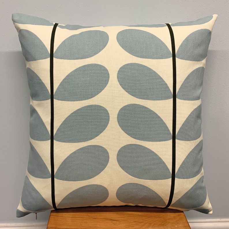 May include: Square throw pillow with a light blue teardrop pattern on a cream background. The pillow has black vertical stripes and a zipper closure. The pillow is placed on a wooden surface.