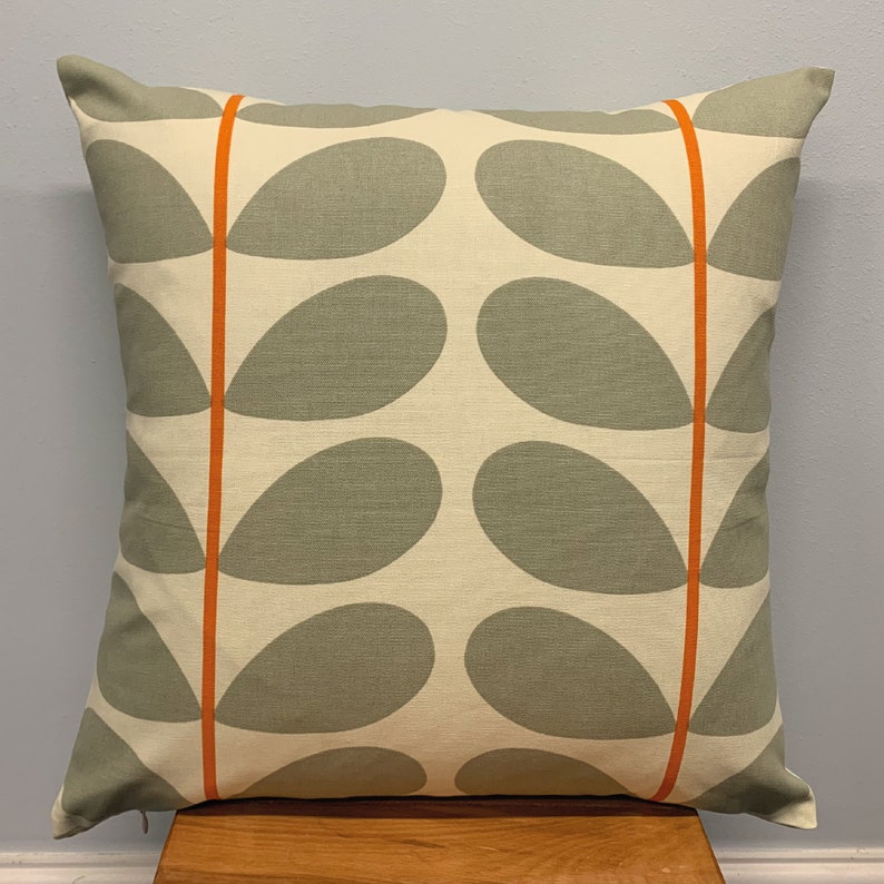 May include: Square decorative pillow with a cream background and a repeating pattern of gray leaf shapes. Two vertical orange stripes run down the pillow's face. The pillow is placed on a wooden surface.
