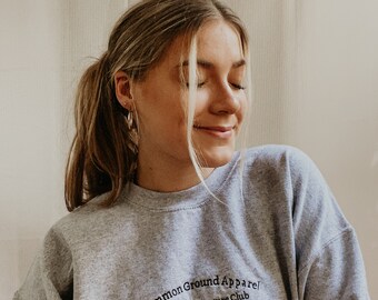 Op de afbeelding: Een grijs sweatshirt met de tekst "Common Ground Apparel Hiking & Adventure Club est. 2022" geborduurd op de voorkant.