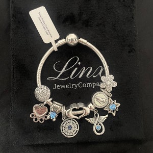 May include: Silver snake chain bracelet with charms, including a heart with the words "You are my heart", a compass, a hand, a dove, a star, and a flower. The bracelet is on a black velvet background.