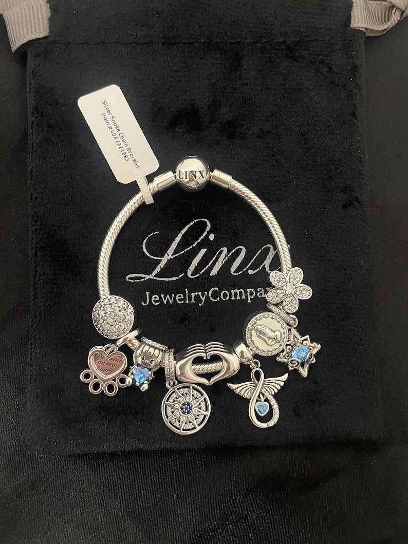 May include: Silver snake chain bracelet with charms, including a heart with the words "You are my heart", a compass, a hand, a dove, a star, and a flower. The bracelet is on a black velvet background.