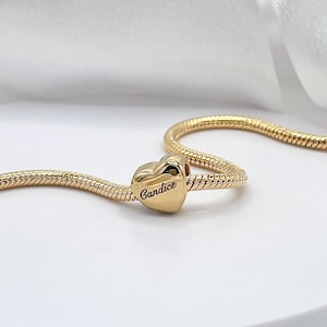 May include: A gold-colored heart-shaped charm with the name "Candice" engraved on it. The charm is on a gold snake chain, displayed on a white surface. The charm is a jewelry item.