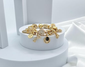 May include: A gold charm bracelet featuring a selection of charms, including a heart, a bird, a tree, and a circle adorned with rhinestones. The bracelet is displayed on a white stand.