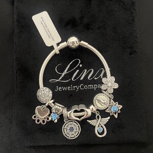 May include: Silver snake chain bracelet with charms, including a heart charm with the inscription "Forever in my heart", a compass charm, a wing charm, a flower charm, and a blue crystal charm. The bracelet is on a black velvet background.