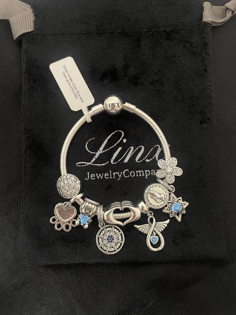 May include: Silver snake chain bracelet with charms, including a heart charm with the inscription "Forever in my heart", a compass charm, a wing charm, a flower charm, and a blue crystal charm. The bracelet is on a black velvet background.