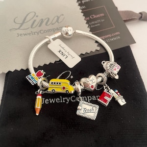 Puede incluir: Un brazalete de plata con una variedad de dijes, incluyendo un autobús escolar, un lápiz, un libro, un corazón con las palabras "Enseñar es amar", y un pequeño libro rojo con una estrella. El brazalete está sobre un fondo de terciopelo negro.