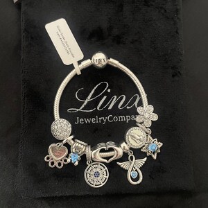 May include: Silver Snake Chain Bracelet with charms, including a paw print charm with the text "You're always on my heart", a compass charm, a hand charm, a wing charm with an infinity symbol, a star charm, and a flower charm. The bracelet is in a black velvet pouch with a tag that reads "Silver Snake Chain Bracelet Item # 520721593".