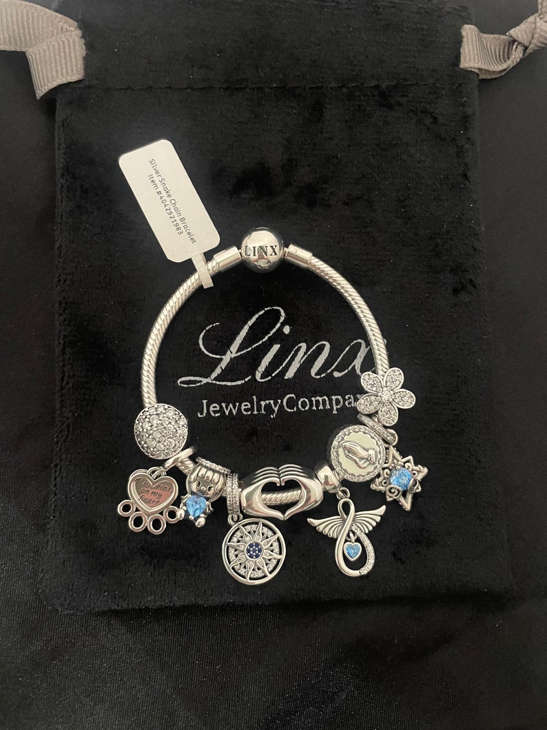 May include: Silver Snake Chain Bracelet with charms, including a paw print charm with the text "You're always on my heart", a compass charm, a hand charm, a wing charm with an infinity symbol, a star charm, and a flower charm. The bracelet is in a black velvet pouch with a tag that reads "Silver Snake Chain Bracelet Item # 520721593".