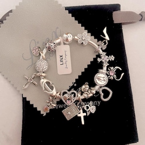 May include: Silver charm bracelet with a variety of charms, including hearts, crosses, a teddy bear, a flower, a religious figure, and a pink stone. The bracelet is on a black background with a small white tag that says "LINX Jewelry Company".