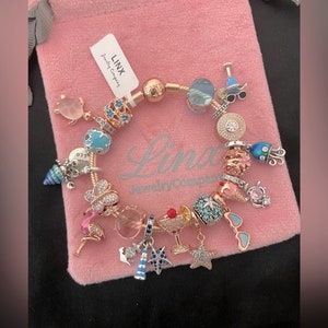 May include: A rose gold charm bracelet with various charms, including a flamingo, a seashell, a starfish, and a cocktail glass. The bracelet is on a pink velvet pouch with the text "Linx Jewelry Company".