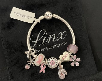 Op de afbeelding: Een zilveren slangenkettingarmband met bedels, waaronder een roze lint, een hartbedel met de tekst "Hope", een bloemenbedel en een wantbedel. De armband ligt op een zwarte fluwelen achtergrond. De tekst op het label luidt "Silver Snake Chain Bracelet Item # 4042921983".