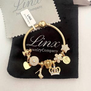 May include: A gold-colored charm bracelet with various charms, including a heart, high heel, crown, star, and flower. The bracelet is displayed on a black pouch with a tag that reads "Linx Jewelry Company."
