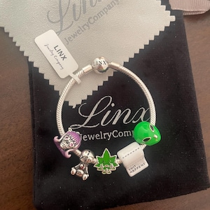 May include: A silver charm bracelet featuring a selection of charms, including a skull, a doll, a marijuana leaf, a toilet roll with the text "SHIT HAPPENS", and an alien. The bracelet is displayed on a black velvet background.