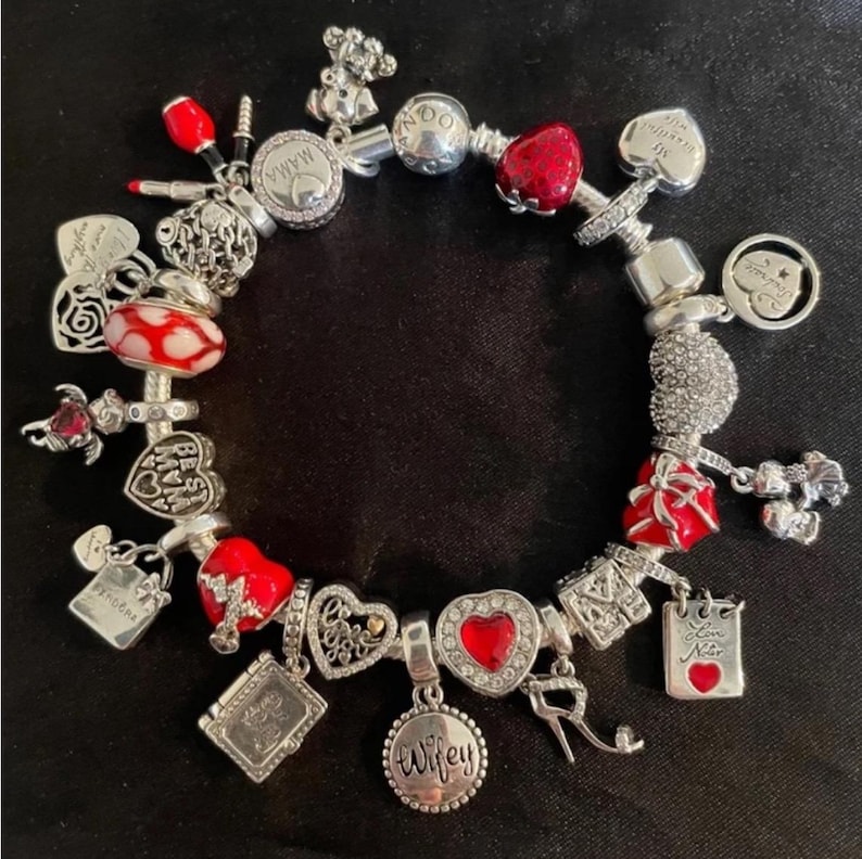 Puede incluir: Un brazalete de plata con dijes rojos y blancos. Los dijes incluyen corazones, una caja de regalo, un l&aacute;piz labial, una rosa y un dije que dice "Wifey".