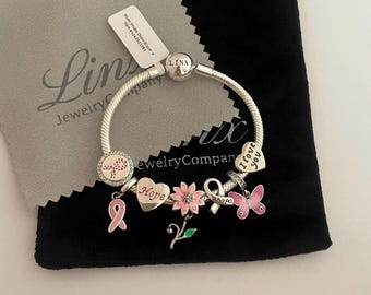 May include: A silver bracelet with pink charms, including a cross, a ribbon, a flower, a butterfly, and a heart with the words "I love you". The bracelet is displayed on a black pouch with a grey cleaning cloth.