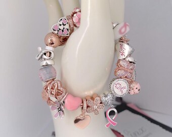 Op de afbeelding: Een armband met roze en zilveren bedels, waaronder harten, vlinders, bloemen en een lint. De armband heeft ook een bedel met het woord "Hope" erop.