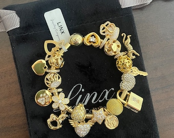 May include: A gold charm bracelet featuring a range of charms, including hearts, stars, a lock, a crown, a paw print, a flower, a horse, and a key. The bracelet is presented in a black velvet pouch with a tag that reads "Linx Jewellery Company".
