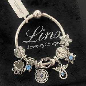 May include: Silver Snake Chain Bracelet with charms including a heart with the inscription "Touch him of my heart", a compass, a praying hands charm, a flower charm, a star charm, a charm with an infinity symbol and a blue crystal charm. The bracelet is on a black velvet background. The bracelet is from Linx Jewelry Company.
