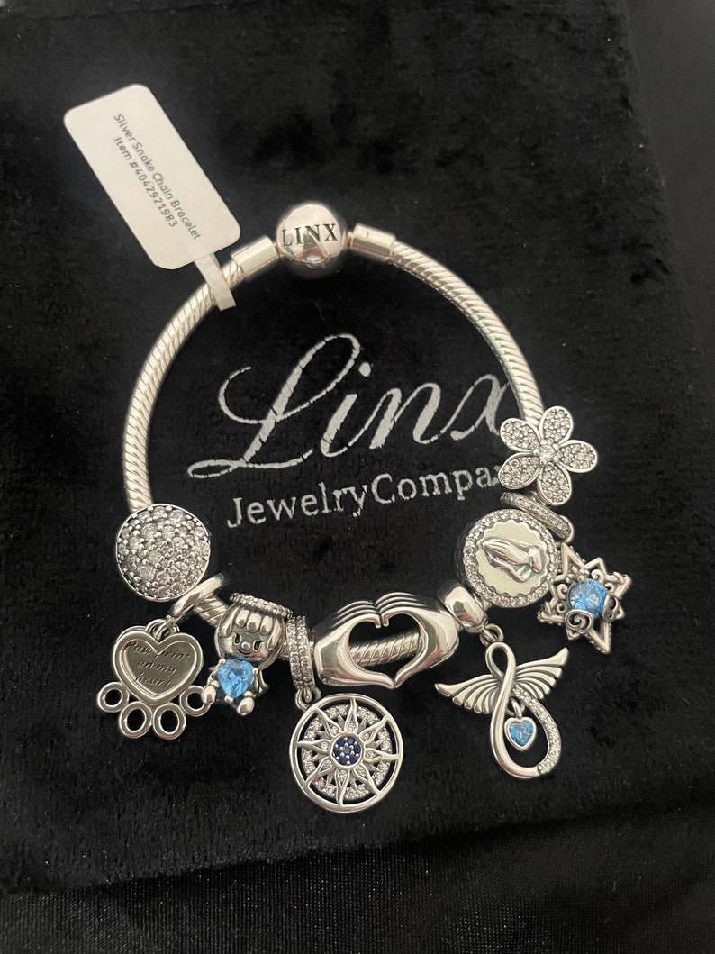 May include: Silver Snake Chain Bracelet with charms including a heart with the inscription "Touch him of my heart", a compass, a praying hands charm, a flower charm, a star charm, a charm with an infinity symbol and a blue crystal charm. The bracelet is on a black velvet background. The bracelet is from Linx Jewelry Company.