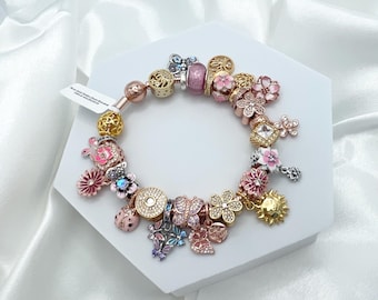 May include: A charm bracelet with a variety of charms, including flowers, butterflies, ladybugs, and a sun. The charms are made of gold, silver, and pink metal. The bracelet is on a white background.