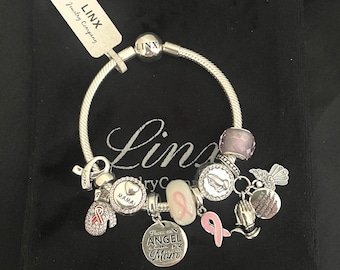 Op de afbeelding: Zilveren armband met roze lintbedels, een hartbedel met de tekst "Mama", een bedel met de tekst "There's an Angel in Heaven My Mom" en een bedel met een gebedshoudende handen ontwerp. De armband ligt op een zwarte fluwelen achtergrond.