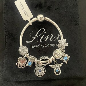 May include: Silver snake chain bracelet with charms, including a paw print charm with "You walk in my heart" text, a compass charm, a heart charm, a flower charm, a hand charm, a winged infinity charm, and a blue crystal charm. The bracelet is on a black velvet background.