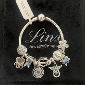 May include: A silver charm bracelet with a variety of charms, including a heart, a paw print, a compass, a flower, a hand, a star, and an angel wing. The bracelet is on a black velvet background. The charms are silver and have blue and white accents. The bracelet is labeled "Linx Jewelry Company".