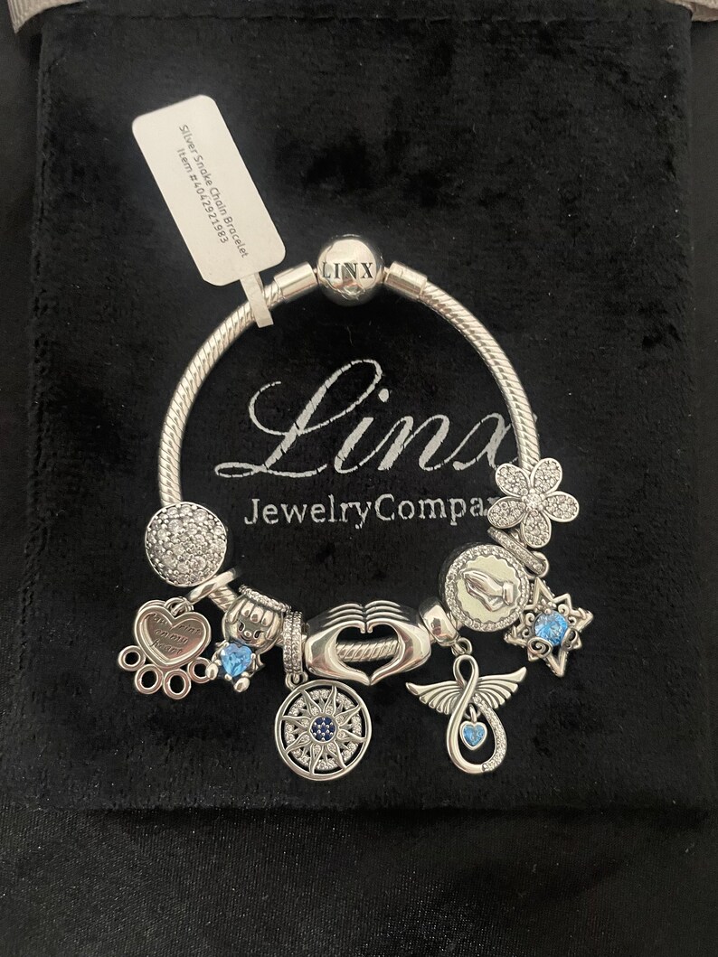 May include: A silver charm bracelet with a variety of charms, including a heart, a paw print, a compass, a flower, a hand, a star, and an angel wing. The bracelet is on a black velvet background. The charms are silver and have blue and white accents. The bracelet is labeled "Linx Jewelry Company".