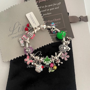 May include: A silver charm bracelet featuring a range of colourful charms, including a cupcake, a skull, a butterfly, a green alien, and a red truck. The bracelet is displayed on a black velvet pouch with the text "Linx Jewelry Company" on it.
