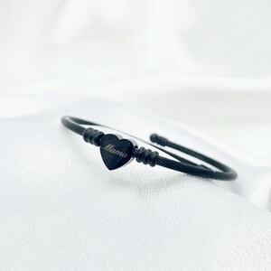 May include: Black cuff bracelet with a heart-shaped charm. The charm has the word "Mama" engraved on it. The bracelet is made of a twisted metal cable and is displayed on a white surface.