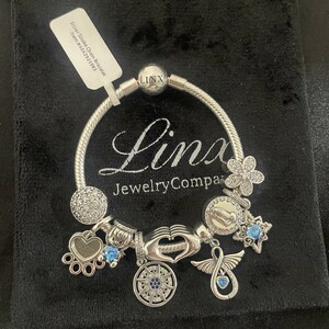 May include: Silver Snake Chain Bracelet with charms including a heart, infinity symbol, paw print, compass, and flower. The bracelet is on a black velvet background. The text "Linx JewelryCompa" is visible on the bracelet.
