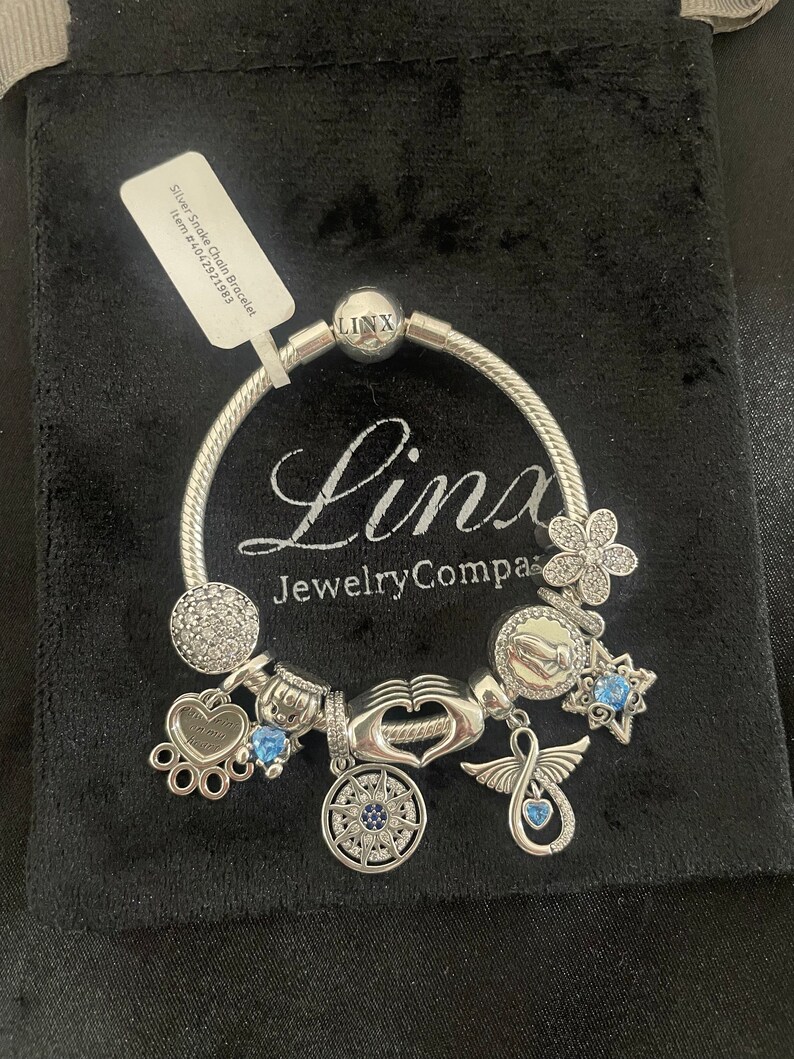 May include: Silver Snake Chain Bracelet with charms including a heart, infinity symbol, paw print, compass, and flower. The bracelet is on a black velvet background. The text "Linx JewelryCompa" is visible on the bracelet.