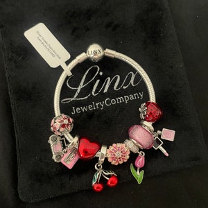 May include: A silver snake chain bracelet with charms, including a red heart charm with the text "You are loved", a pink flower charm, a red and white strawberry charm, a pink and white charm with a floral design, a pink and white charm with a radio design, a pink and white charm with a lipstick design, a pink and white charm with a tulip design, and a red cherry charm. The bracelet is on a black velvet background. The text "Linx Jewelry Company" is visible on the bracelet.
