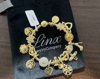 May include: A gold charm bracelet featuring a range of charms, including a heart, a bee, a tree, a paw print, a sun, a flower, a crown, a shoe, and a bee. The bracelet is displayed on a black velvet pouch with the text "Linx Jewellery Company" on a white tag.