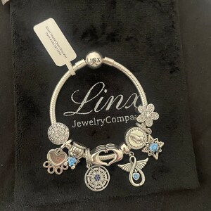 May include: Silver Snake Chain Bracelet with charms including a compass, a heart with wings, a hand with a heart, a paw print, a star, a flower, and a praying hands charm. The bracelet is on a black velvet background.  The text on the tag reads "Silver Snake Chain Bracelet Item # 64027521583".