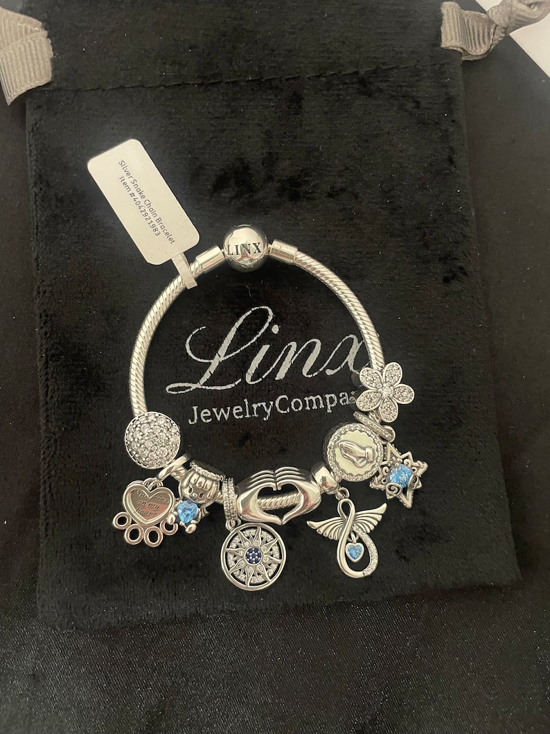 May include: Silver Snake Chain Bracelet with charms including a compass, a heart with wings, a hand with a heart, a paw print, a star, a flower, and a praying hands charm. The bracelet is on a black velvet background.  The text on the tag reads "Silver Snake Chain Bracelet Item # 64027521583".