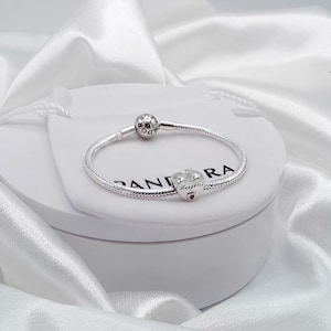 Puede incluir: Una pulsera de cadena de serpiente plateada con un dije en forma de corazón grabado con la palabra Daughter y una pequeña piedra roja. La pulsera incluye un cierre redondo con el nombre de la marca Pandora grabado en la superficie del metal.
